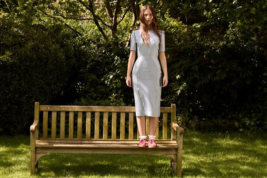 Woman in a light dress standing on a wooden bench in a garden with trees and grass.